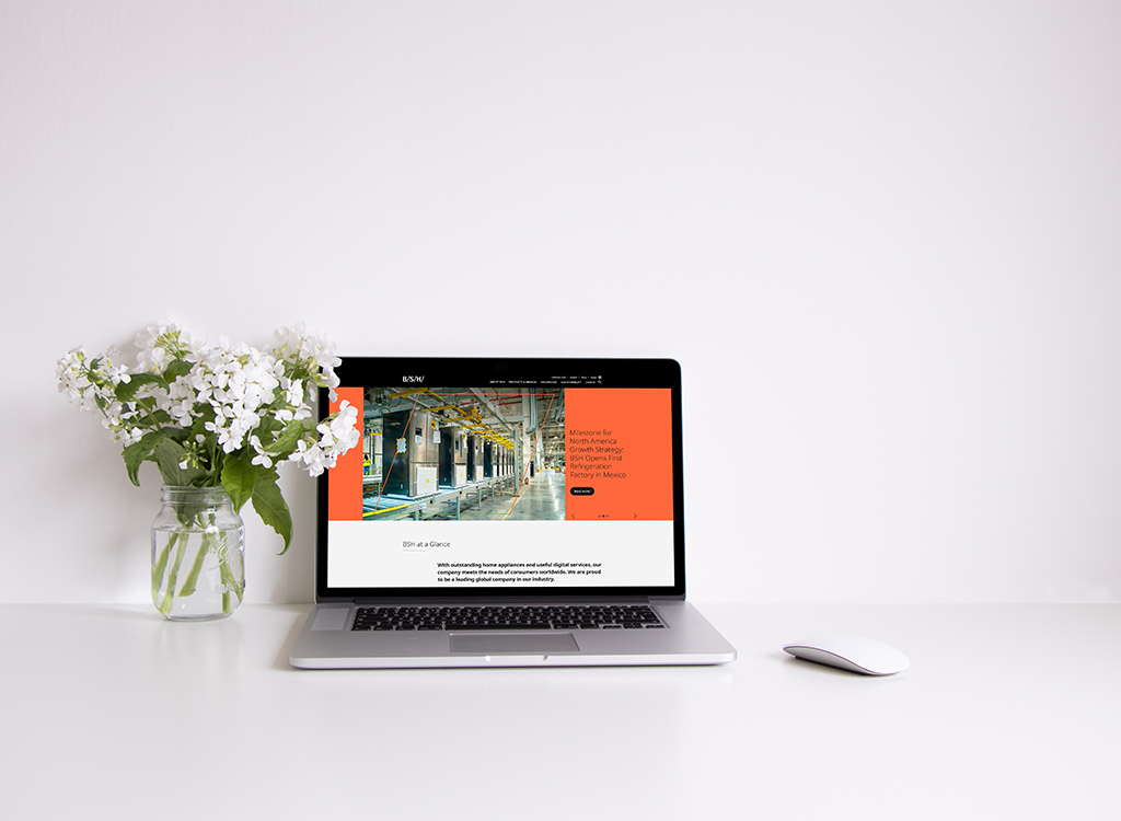 A modern laptop displaying a website on a minimalist desk with a white background, featuring a vase of white flowers and a sleek mouse, emphasizing a clean and professional workspace aesthetic.