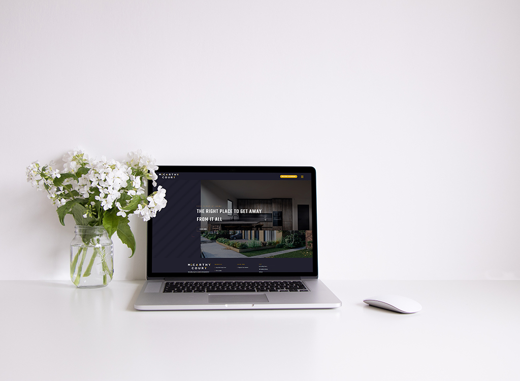 A modern laptop displaying a real estate website sits on a clean, minimalistic desk alongside a vase of white flowers and a sleek mouse, creating a stylish workspace atmosphere.