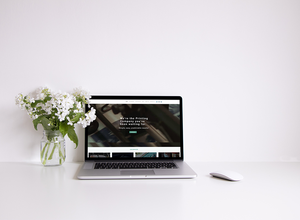 A modern workspace featuring a laptop displaying a printing company website, accompanied by a vase of white flowers, on a clean white desk.