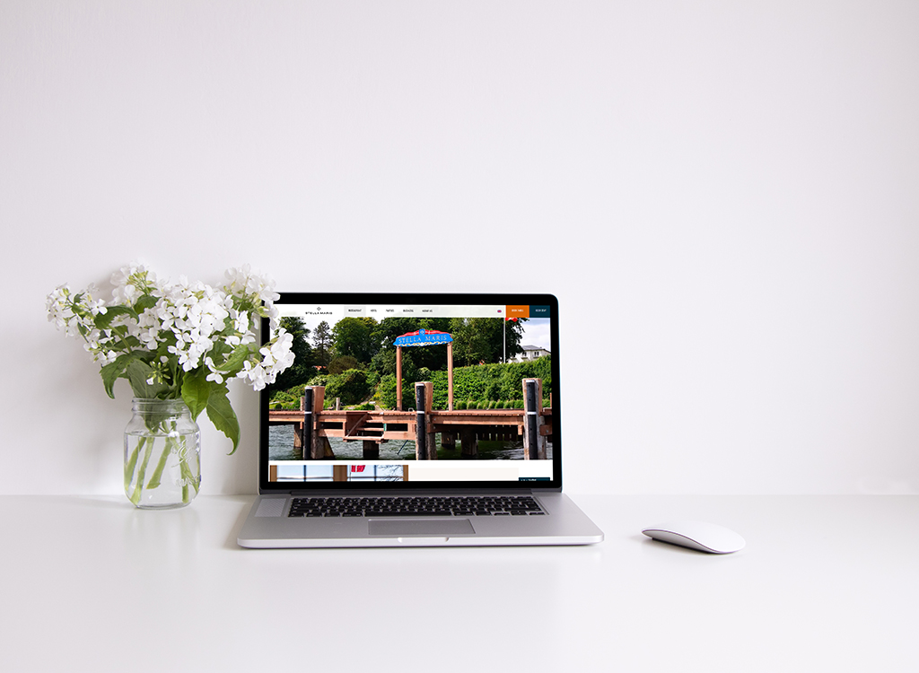 Laptop displaying a website with a dock and greenery, placed on a clean white desk next to a vase of white flowers.