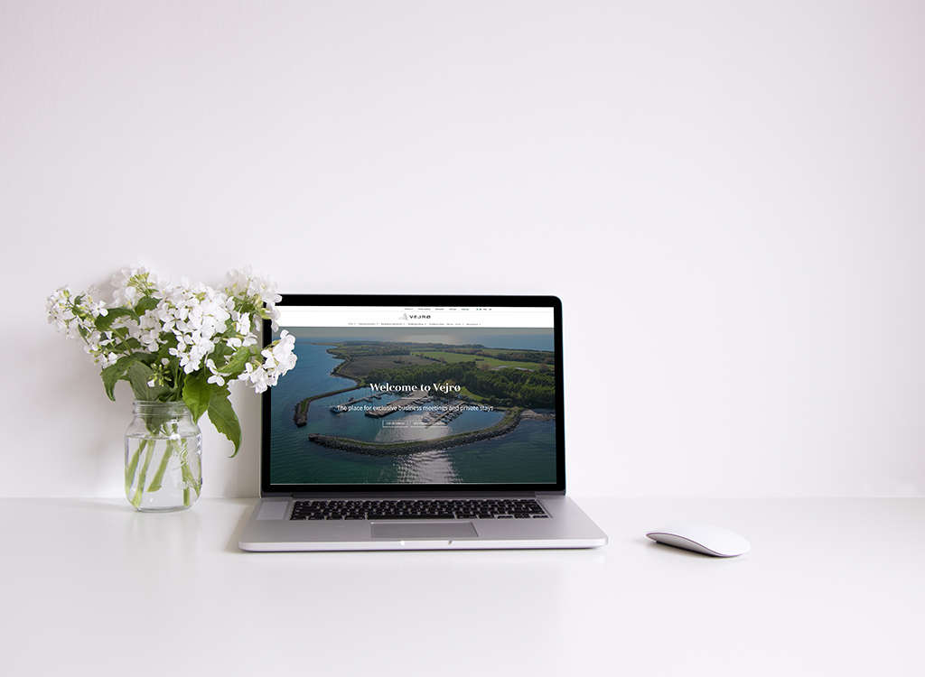 Laptop displaying the website "Welcome to Vejro," showcasing a scenic aerial view of a coastal area, accompanied by a vase of white flowers on a minimalist desk.